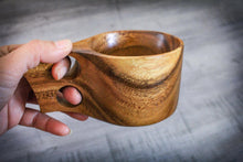 Load image into Gallery viewer, Hand hält einen handgefertigten Kuksa-Becher aus Akazienholz im skandinavischen Design; gut sichtbare ergonomische Doppelgriff-Form und natürliche Holzmaserung vor grauem Hintergrund.
