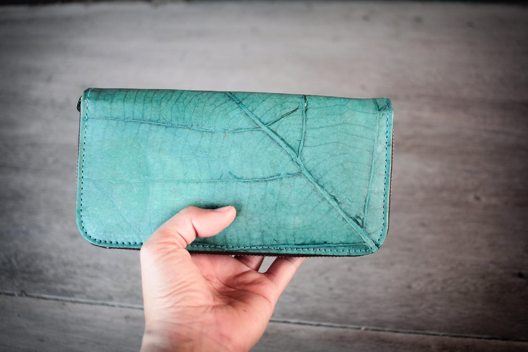Eine Hand hält eine nachhaltige, vegane Brieftasche in Türkis in die Kamera. Die Geldbörse besteht aus recycelten Blättern und zeigt eine deutliche Blattrippen-Struktur.

A hand holding a sustainable, vegan turquoise wallet toward the camera. The wallet is made from recycled leaves and clearly shows the distinct leaf vein structure.