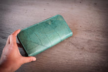 Laden des Bilds im Galerie-Viewer, Eine Hand hält eine handgefertigte, vegane Geldbörse in Türkis, die aus recycelten Blättern hergestellt wurde. Die feine Blattstruktur ist auf dem Material erkennbar.

A hand holding a handmade, vegan turquoise wallet made from recycled leaves. The distinct leaf vein pattern is visible on the surface.
